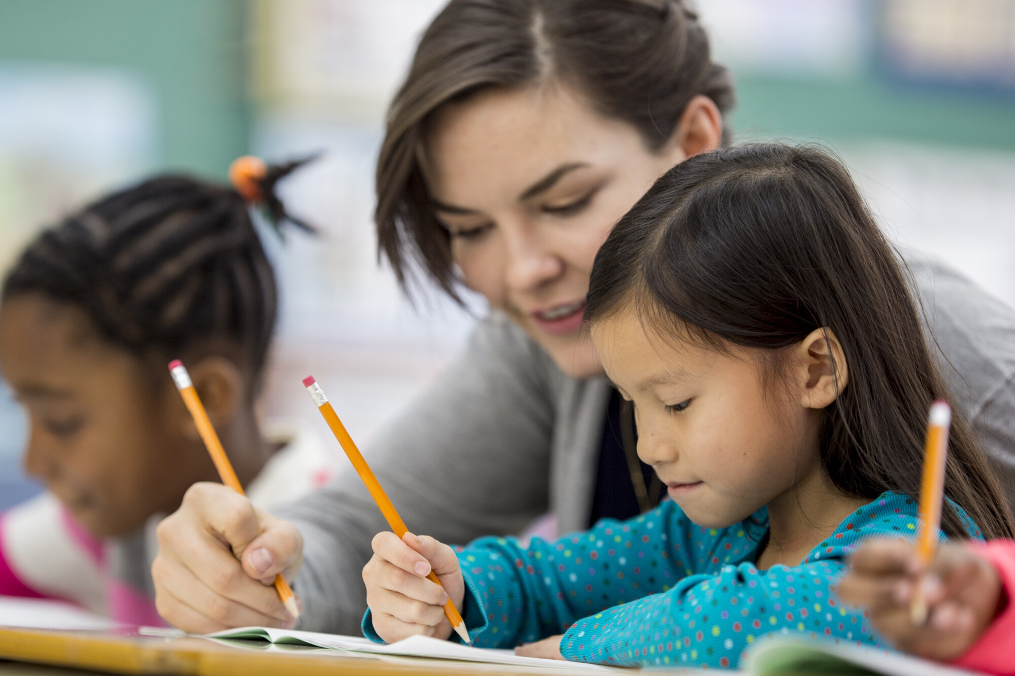 A multi-ethnic group of elementary school children are indoors in a classroom. The students are sitting at their desks and writing with pencils and getting help from a teacher.