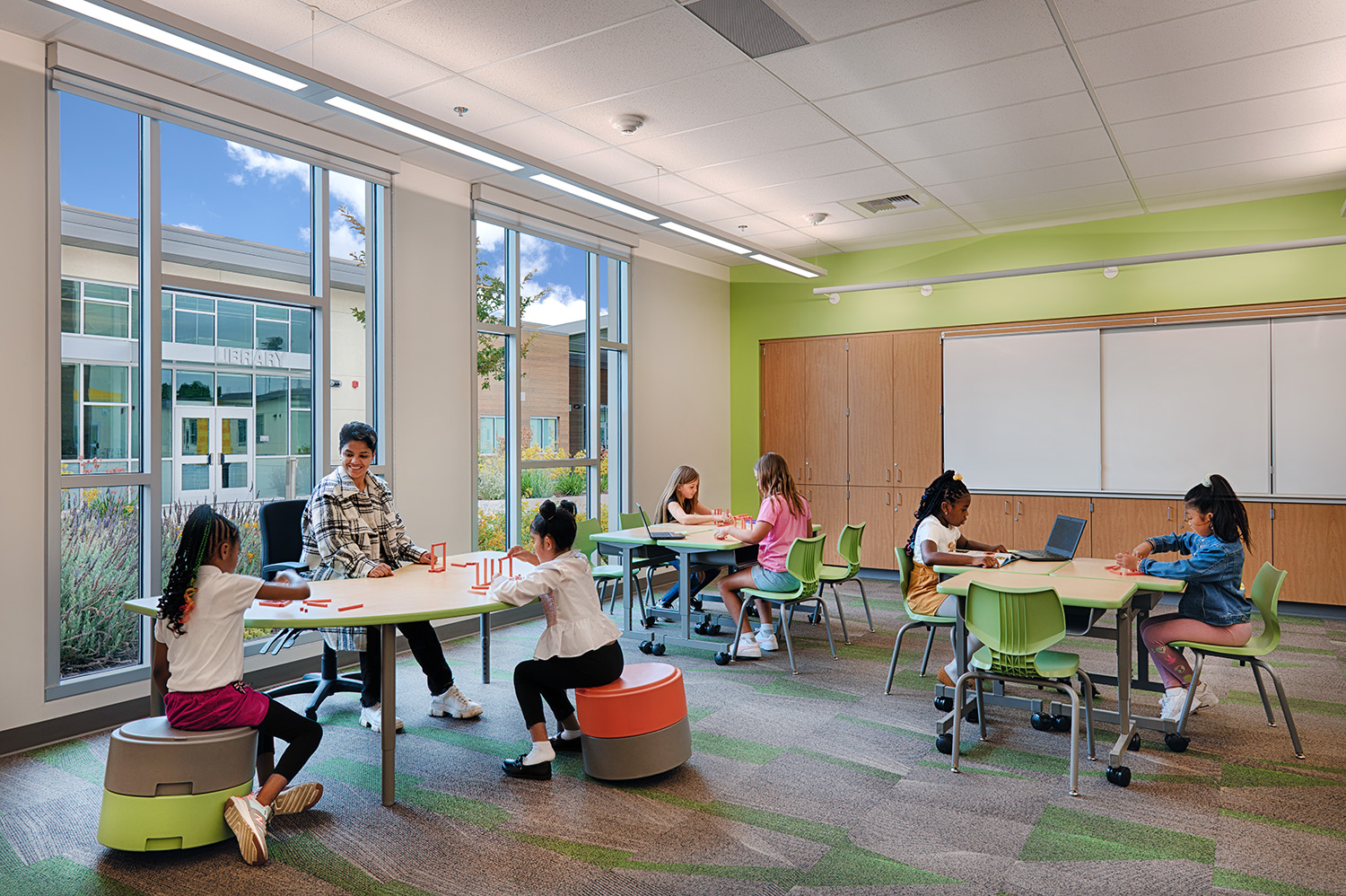 Image of a teacher and students in a colorful, modern classroom with large windows overlooking an adjacent library.